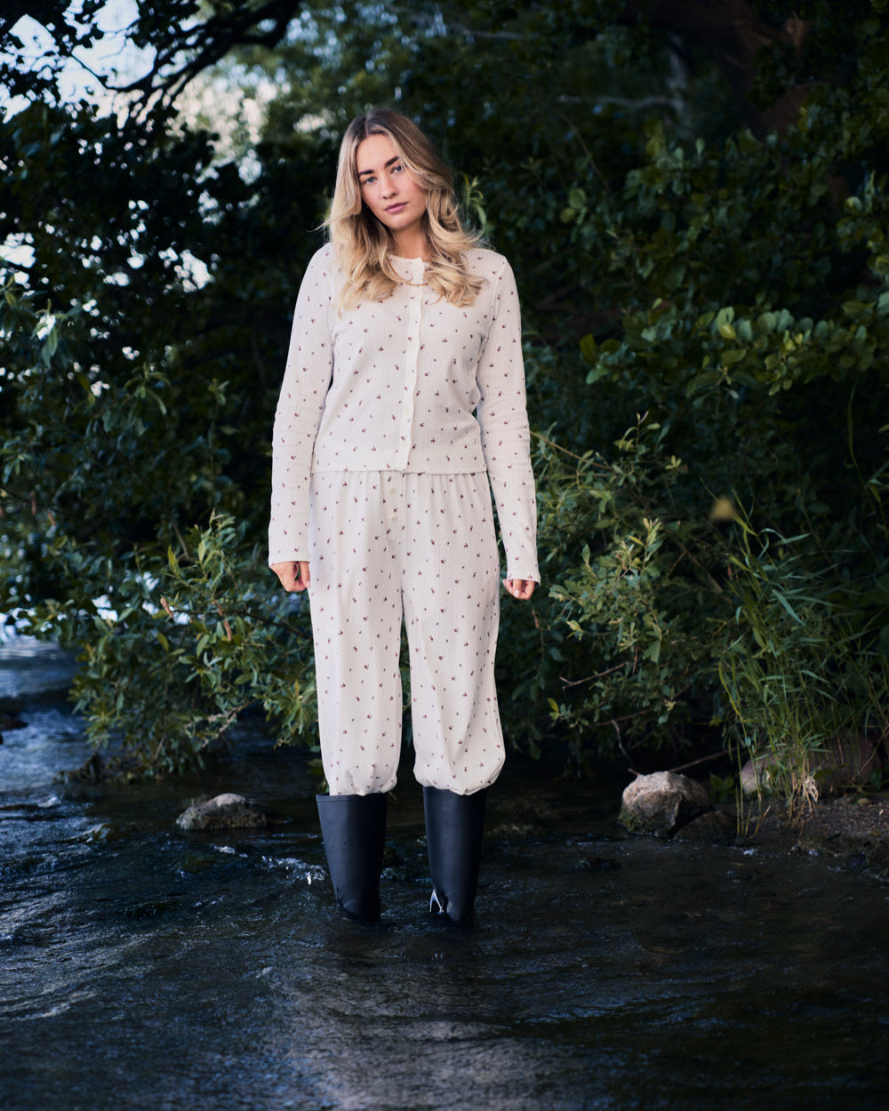 A woman with long blonde hair stands in shallow water, wearing jan-n-june POINTELLE PYJAMA PANTS BLOOMY GOTS and black rubber boots, surrounded by green trees and foliage.