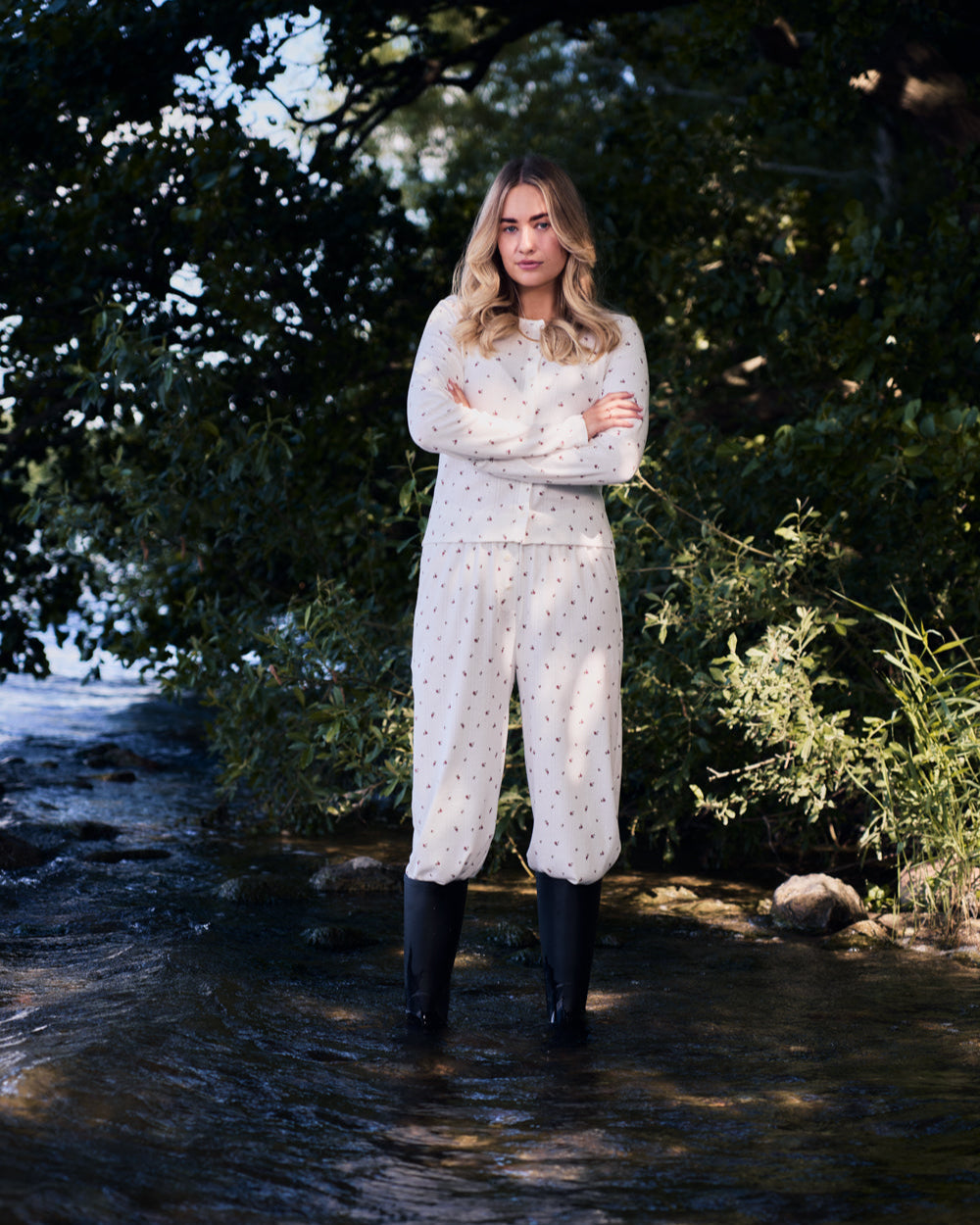 A woman with long blonde hair stands in shallow water, wearing jan-n-junes POINTELLE PYJAMA PANTS BLOOMY GOTS, a cream set with small patterns and pointelle fabric, paired with black rain boots, arms crossed amid lush greenery.