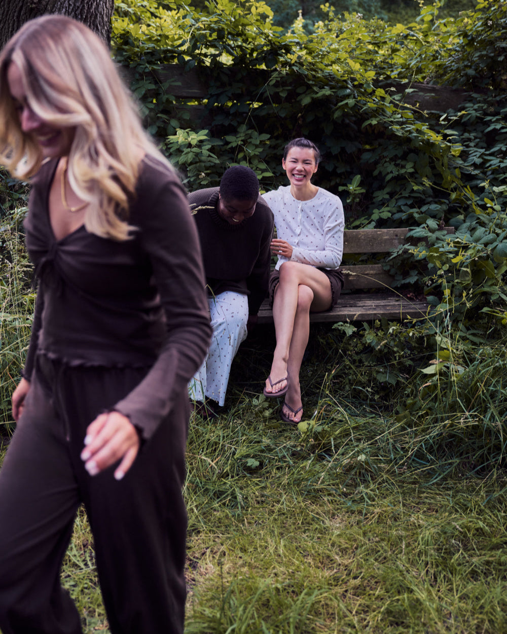 Three women enjoy a park: two laugh on a bench while one walks in front wearing jan-n-junes POINTELLE PYJAMA PANTS CHOCOLATE GOTS. Greenery and overgrown grass surround them, creating a relaxed, natural vibe.