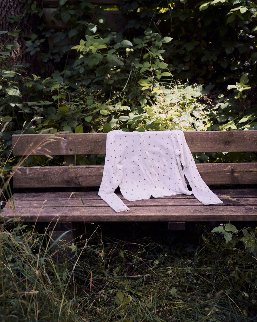 The jan-n-june POINTELLE PYJAMA BUTTONED LONGSLEEVE BLOOMY GOTS, a white organic cotton top with subtle patterns, rests on a weathered wooden bench amid lush grass and leafy plants in a sunlit outdoor setting.