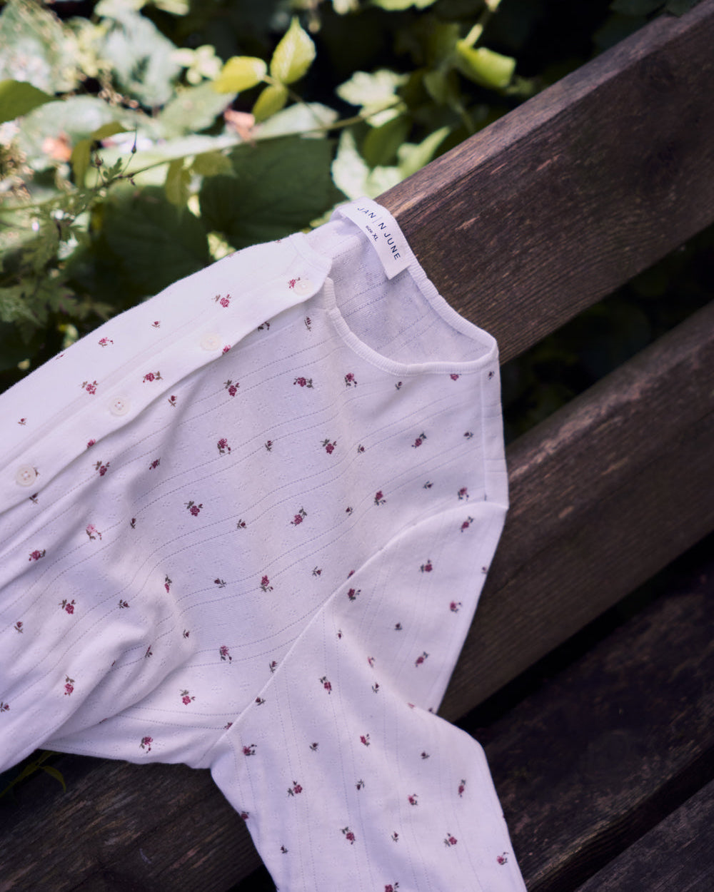 The jan-n-june POINTELLE PYJAMA BUTTONED LONGSLEEVE BLOOMY GOTS, a soft organic cotton white baby shirt with red floral prints, is draped over a wooden bench with green foliage in the background.