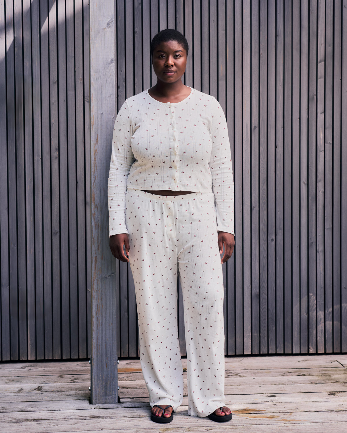 A person stands outdoors on a wooden deck, wearing jan-n-junes POINTELLE PYJAMA PANTS BLOOMY GOTS—white, loose-fitting, organic cotton pants with a subtle dotted pattern—paired with a matching button-up top and black sandals by vertical wooden slats.