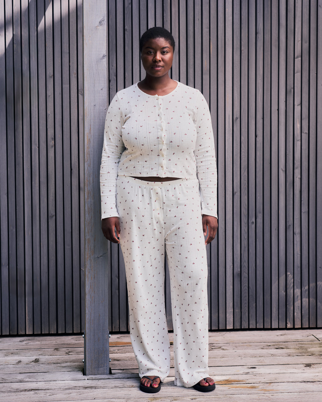 A woman with short hair stands outdoors by wooden panels, wearing the jan-n-june POINTELLE PYJAMA BUTTONED LONGSLEEVE BLOOMY GOTS in cream with a dotted pattern and black sandals.
