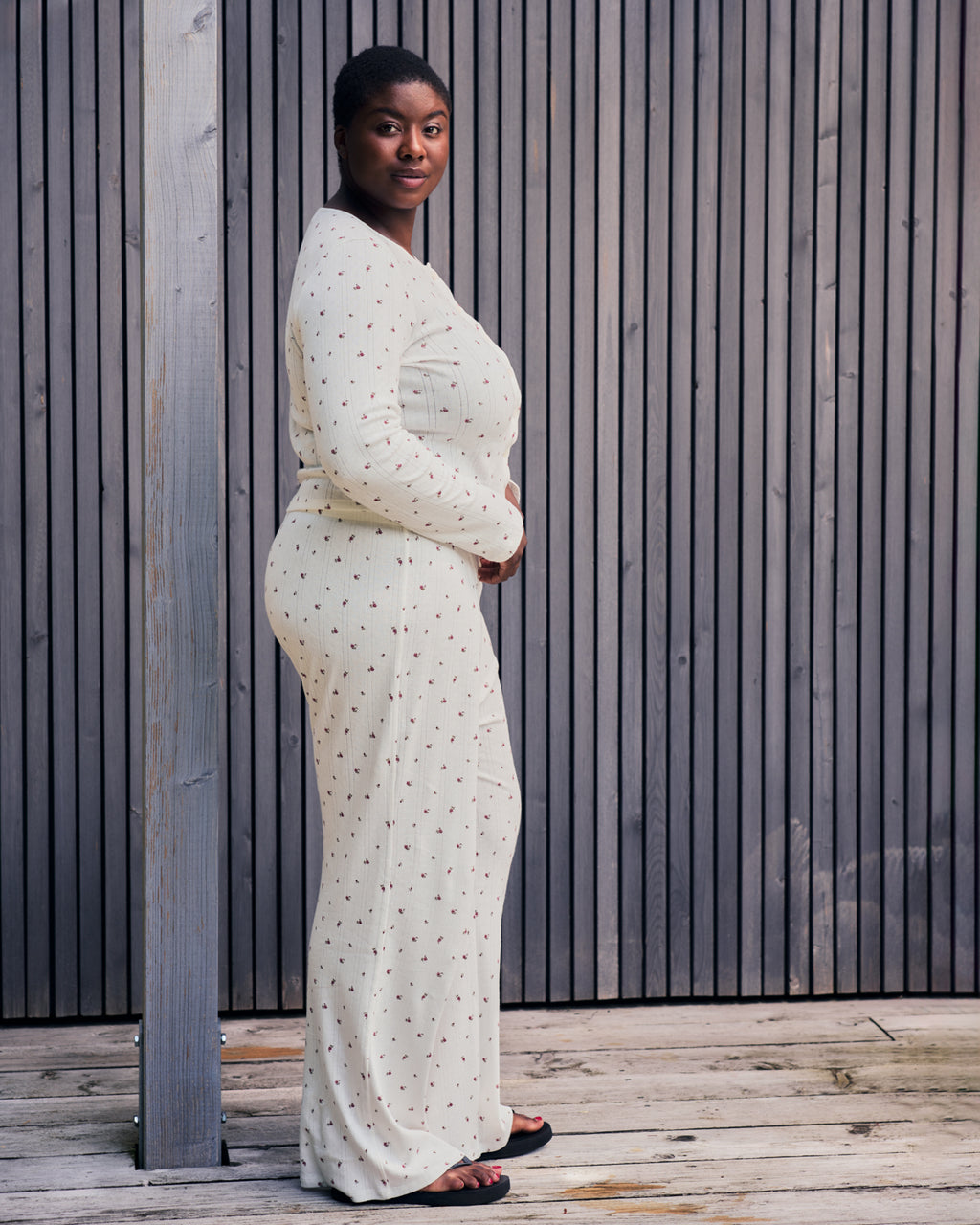 A woman stands sideways, looking at the camera, wearing the jan-n-june POINTELLE PYJAMA BUTTONED LONGSLEEVE BLOOMY GOTS—a long-sleeved, patterned organic cotton dress—on a wooden floor before a modern vertical wooden slat wall.