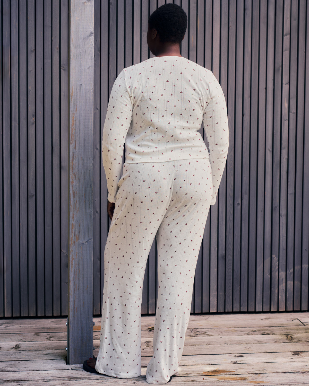 A person stands on a deck by a slatted fence, wearing jan-n-junes POINTELLE PYJAMA PANTS BLOOMY GOTS—a cream, long-sleeved pajama set with subtle dot patterns—paired with black slippers.