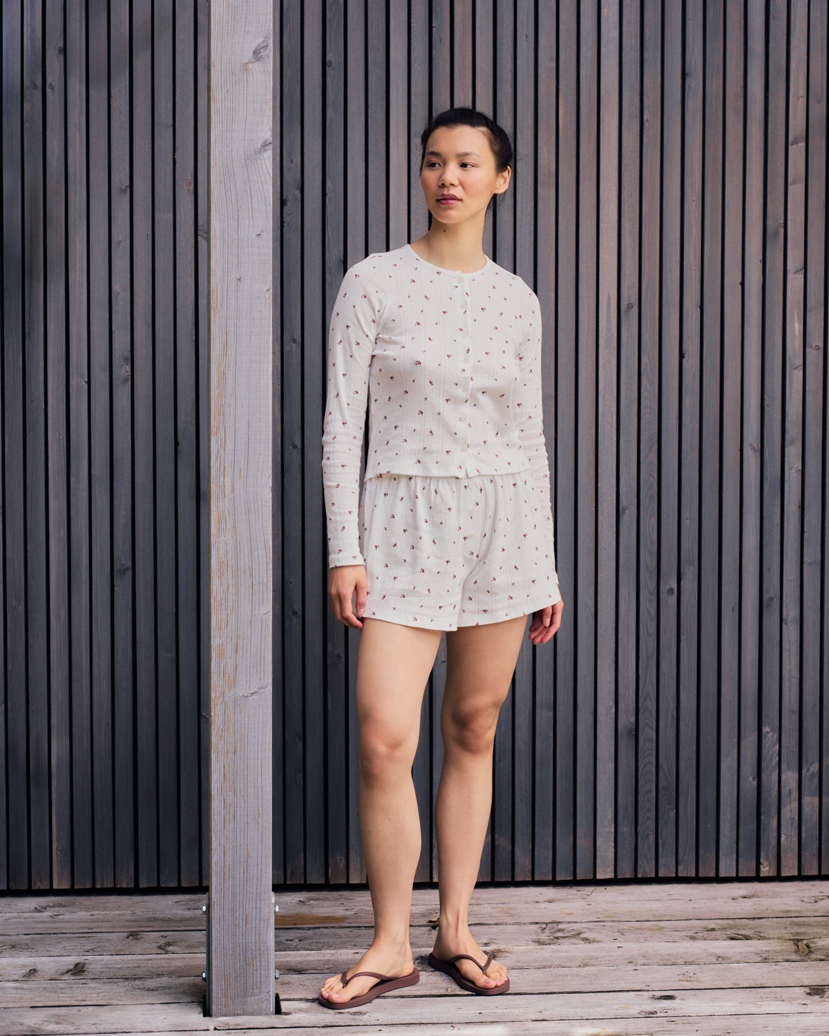 A woman wearing jan-n-june’s POINTELLE PYJAMA SHORTS BLOOMY GOTS and a white long-sleeve top stands on a wooden deck with vertical slats, in flip-flops, gazing to the side.