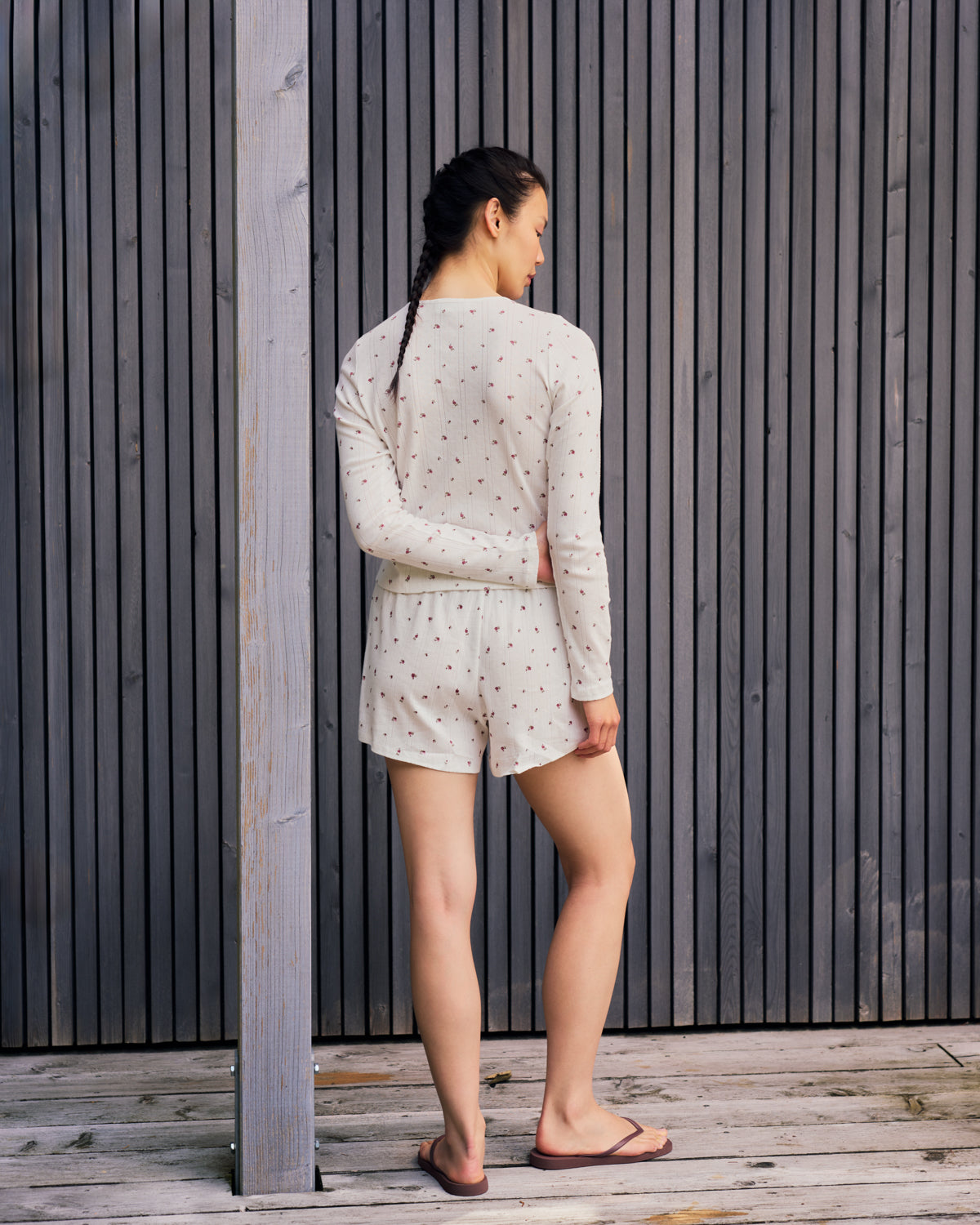 A woman with a braid stands on a wooden deck, facing away, wearing jan-n-junes POINTELLE PYJAMA SHORTS BLOOMY GOTS—light, long-sleeve, polka-dotted organic cotton pyjamas—with flip-flops. Vertical wooden panel wall in the background.