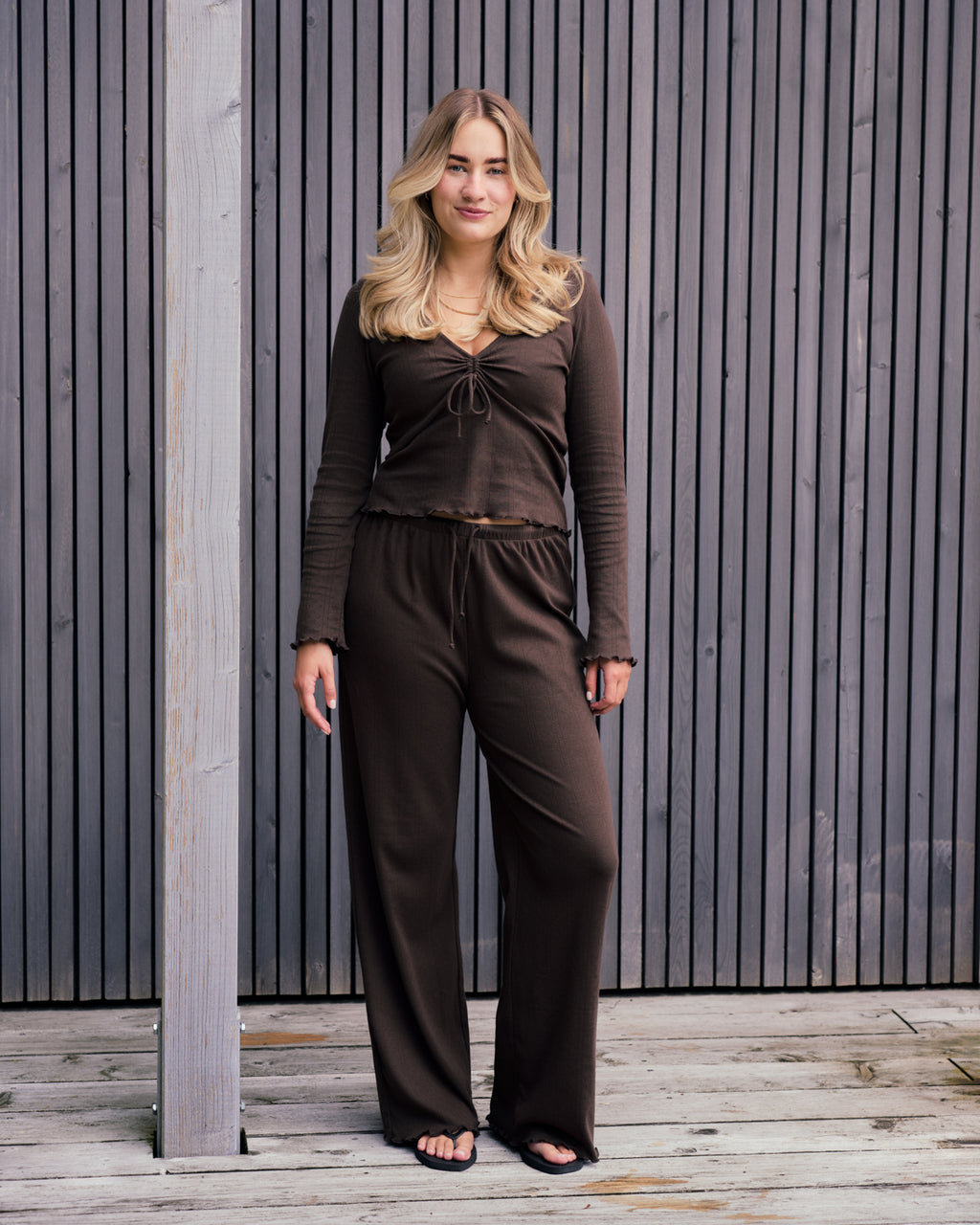 A blonde woman stands on a wooden deck in front of vertical slats, wearing the jan-n-june POINTELLE PYJAMA V-NECK LONGSLEEVE CHOCOLATE GOTS set with wide-legged pants and black sandals.