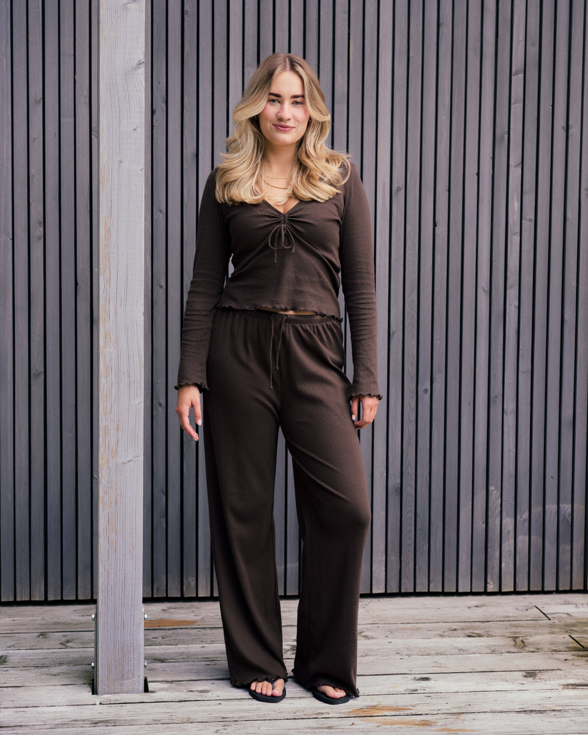 A blonde woman stands on a wooden deck before gray slats, wearing jan-n-june POINTELLE PYJAMA PANTS CHOCOLATE GOTS with a matching top and black sandals, gazing at the camera with a neutral expression.