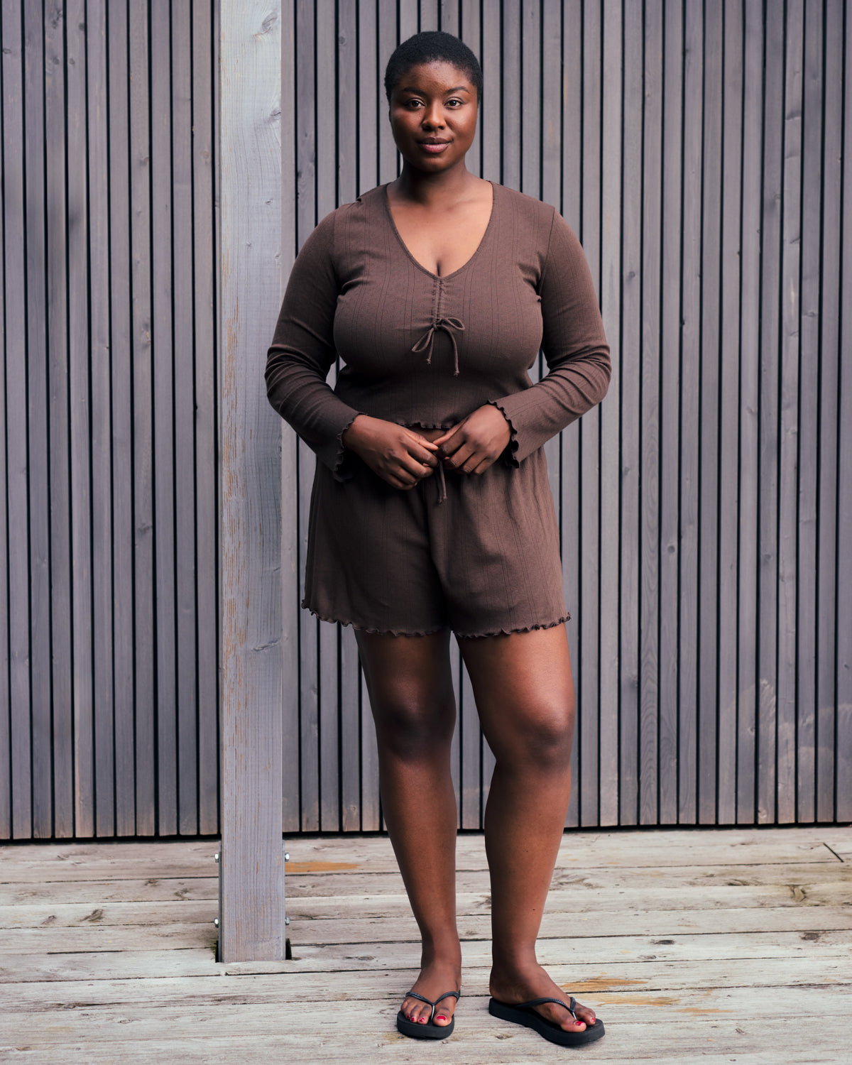 A woman stands on a wooden deck in front of a slat wall, wearing the jan-n-june POINTELLE PYJAMA V-NECK LONGSLEEVE CHOCOLATE GOTS set. She looks at the camera with a relaxed, confident expression.