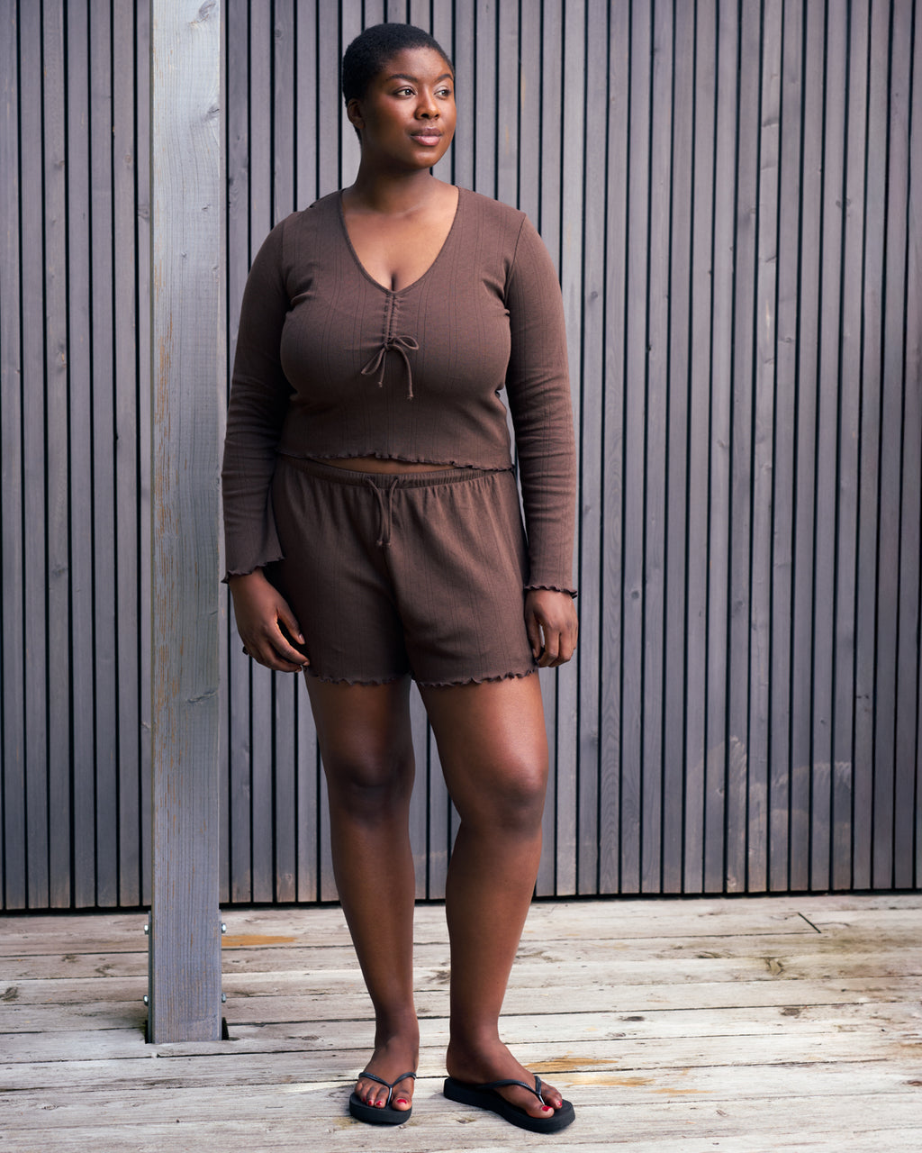 A woman stands on a wooden deck before a slat wall, wearing the jan-n-june POINTELLE PYJAMA V-NECK LONGSLEEVE CHOCOLATE GOTS set with black sandals. She gazes to the side, looking relaxed.