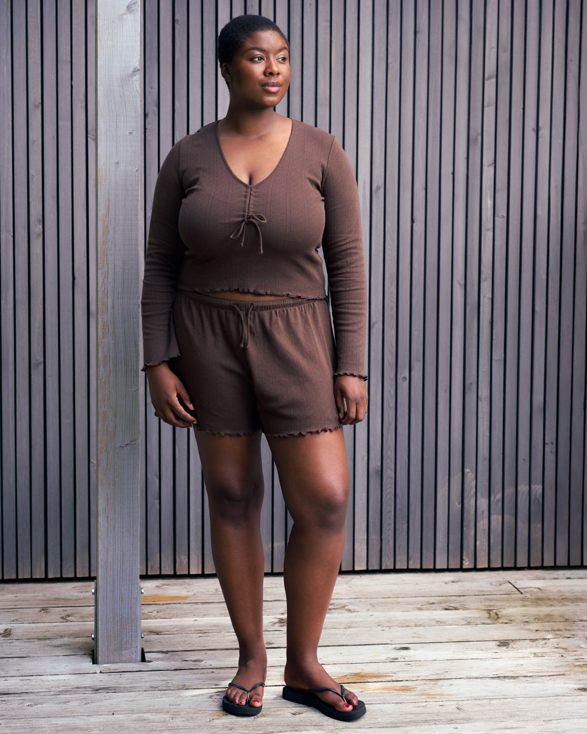 A woman stands on a wooden deck before a slat wall, wearing the jan-n-june POINTELLE PYJAMA V-NECK LONGSLEEVE CHOCOLATE GOTS set with black sandals. She gazes to the side, looking relaxed.
