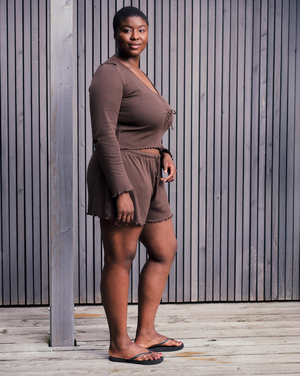 A woman stands on a wooden deck in front of a vertical slat wall, wearing the jan-n-june POINTELLE PYJAMA V-NECK LONGSLEEVE CHOCOLATE GOTS romper and black slide sandals, her expression neutral.