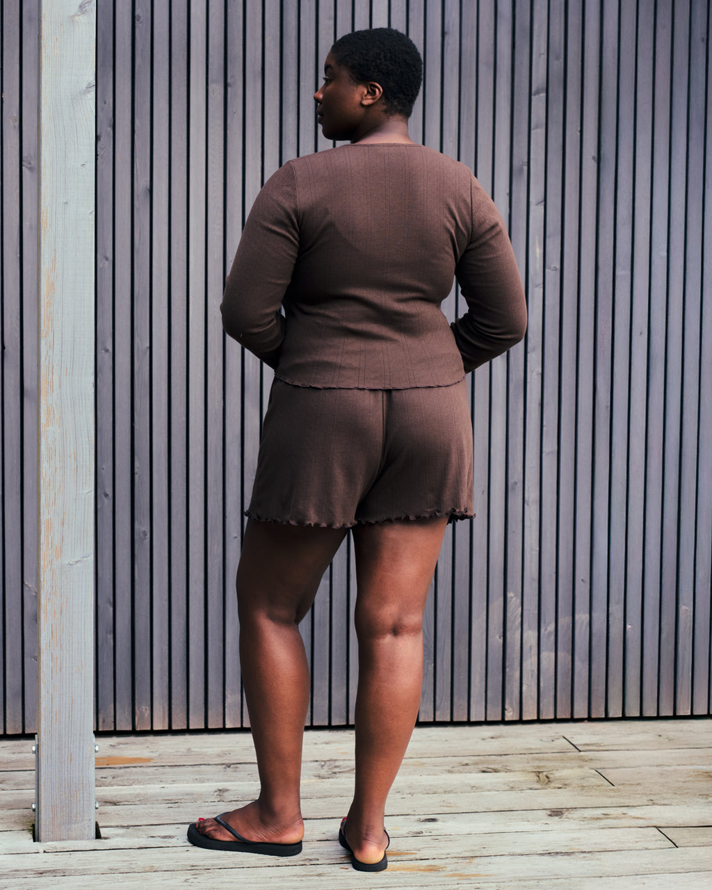 A person stands on a wooden deck in front of a slatted wall, wearing jan-n-june POINTELLE PYJAMA SHORTS CHOCOLATE GOTS and a matching brown top with black sandals.