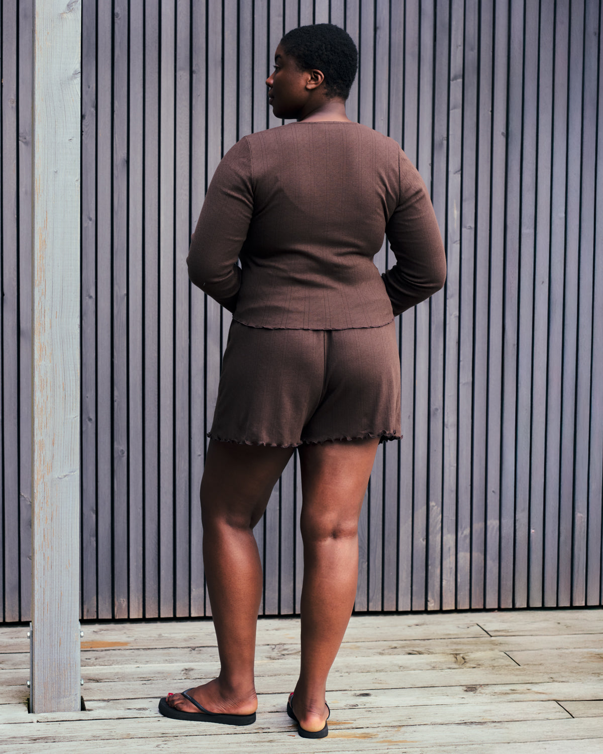 A person stands on a wooden deck in front of a slatted wall, wearing jan-n-june POINTELLE PYJAMA SHORTS CHOCOLATE GOTS and a matching brown top with black sandals.