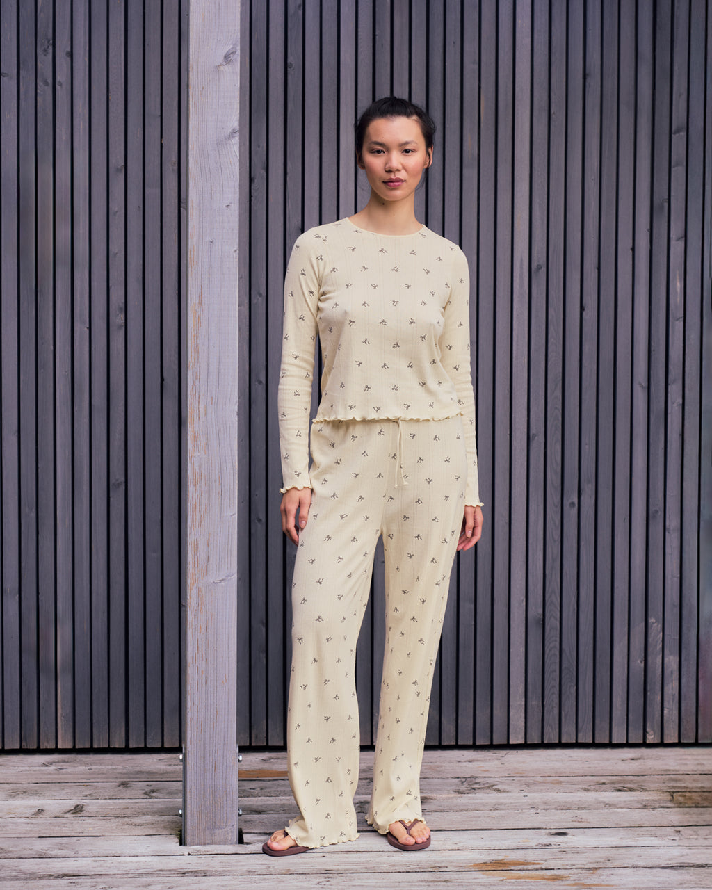 A woman stands on a wooden deck in front of vertical slats, wearing the jan-n-june POINTELLE PYJAMA CREWNECK BUTTERBOW GOTS, a cream-colored organic cotton pyjama set with a small black print, paired with sandals.
