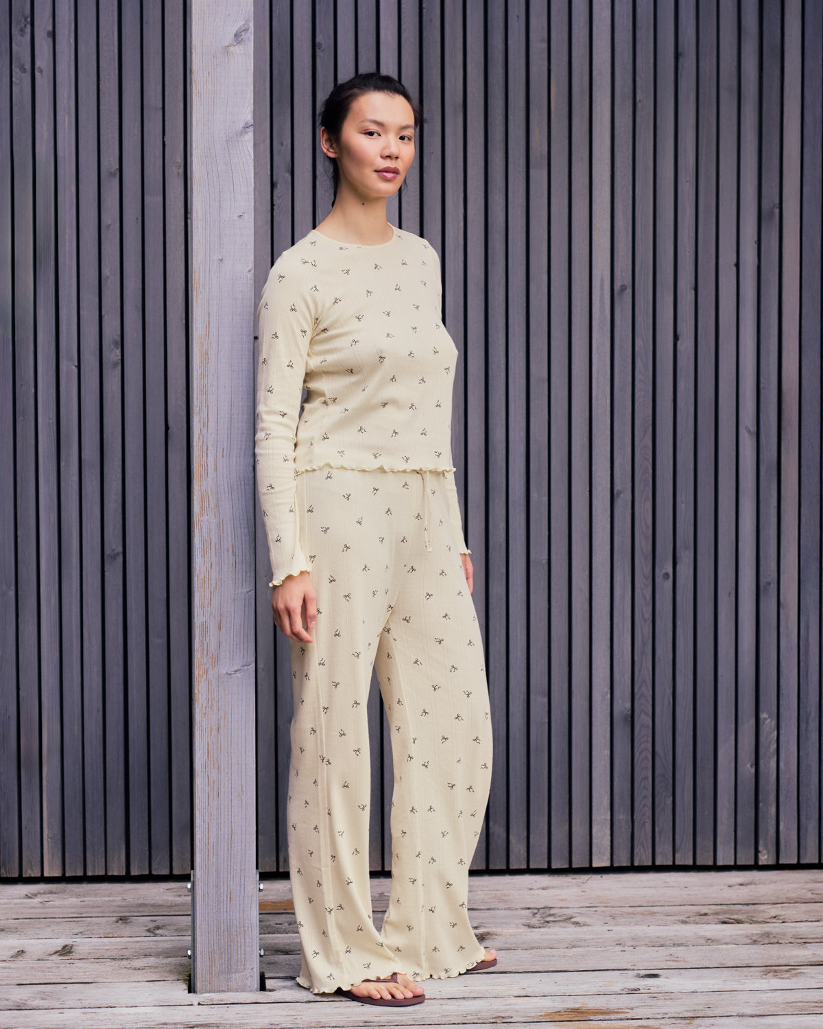 A woman stands outdoors against a wooden slat wall, barefoot and looking ahead, wearing jan-n-junes POINTELLE PYJAMA PANTS BUTTERBOW GOTS, made from light-colored, GOTS-certified organic cotton with a long-sleeve top.
