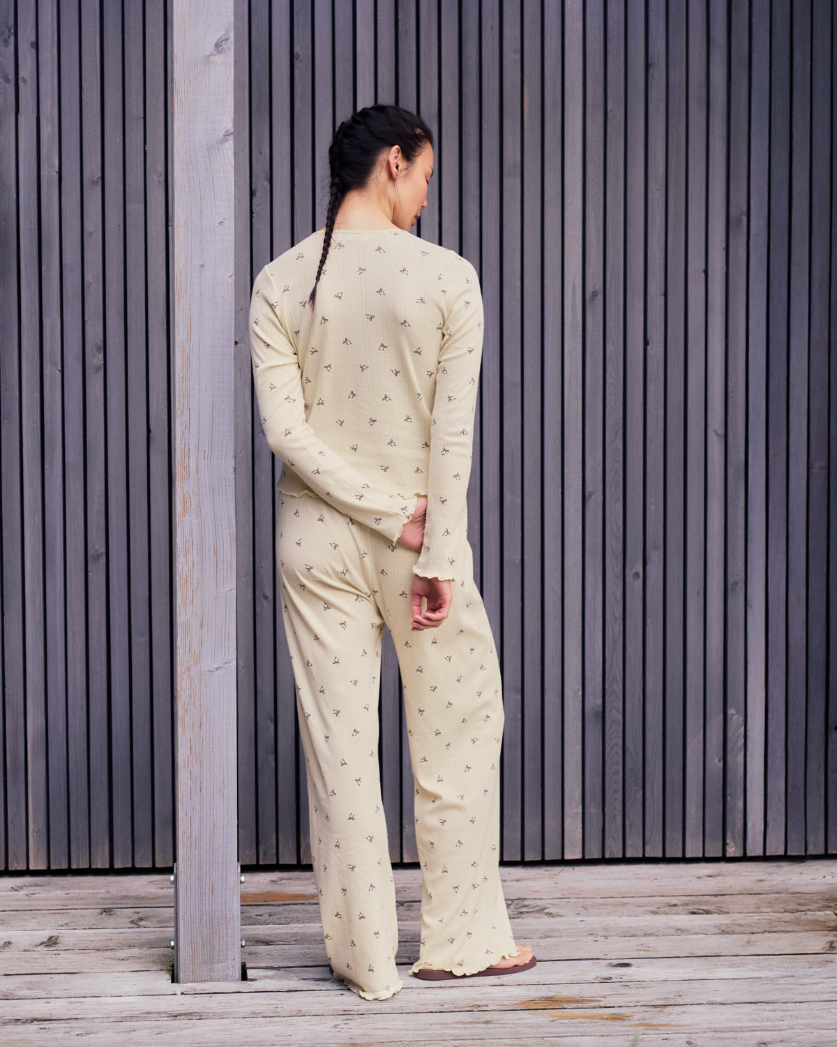 A person with long, dark braids stands facing away, wearing jan-n-june’s POINTELLE PYJAMA PANTS BUTTERBOW GOTS—light yellow, GOTS certified organic cotton with a small black pattern—in front of vertical wooden slats on a wooden deck.