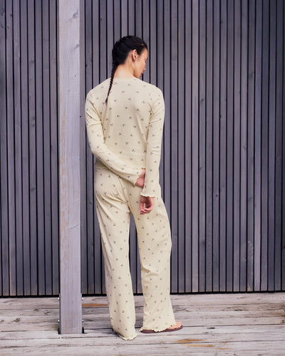 A person with long, dark braids stands facing away, wearing jan-n-june’s POINTELLE PYJAMA PANTS BUTTERBOW GOTS—light yellow, GOTS certified organic cotton with a small black pattern—in front of vertical wooden slats on a wooden deck.