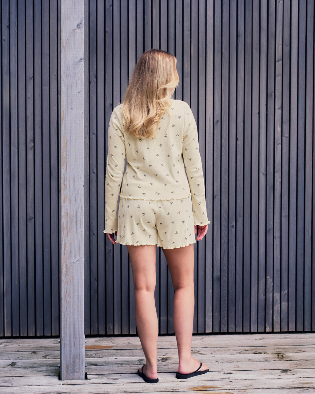 A blonde woman, her back to the camera, faces a dark wooden slatted wall. She wears jan-n-junes POINTELLE PYJAMA SHORTS BUTTERBOW GOTS with a light long-sleeve top and black flip-flops.