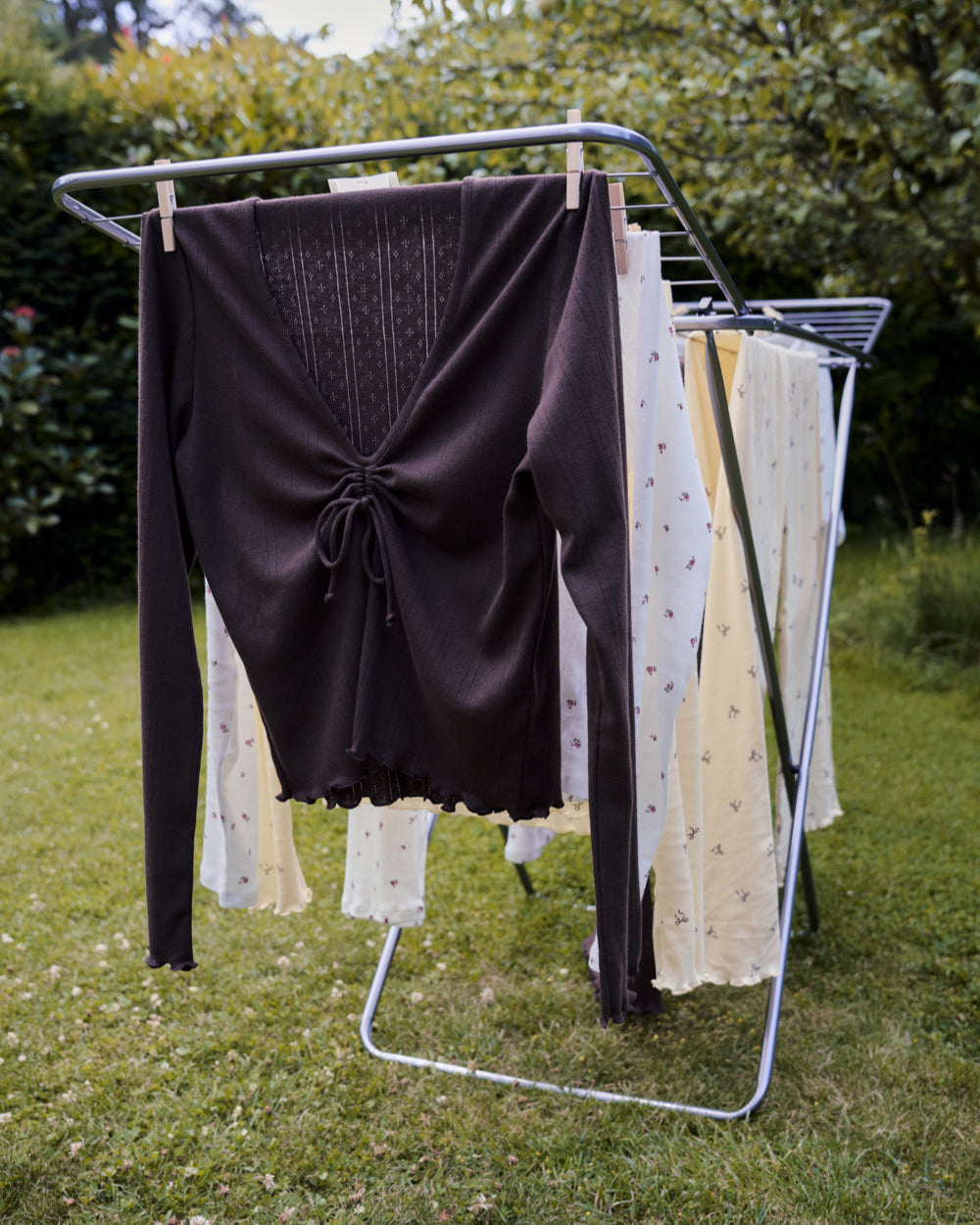 The jan-n-june POINTELLE PYJAMA V-NECK LONGSLEEVE CHOCOLATE GOTS and several light garments hang on a drying rack outdoors on a grassy lawn, surrounded by trees and bushes.