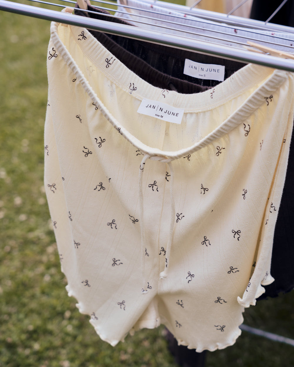 A pair of jan-n-june POINTELLE PYJAMA SHORTS BUTTERBOW GOTS in cream with a small black pattern hangs outdoors on a clothesline, brand label visible on the waistband, with grass in the background.