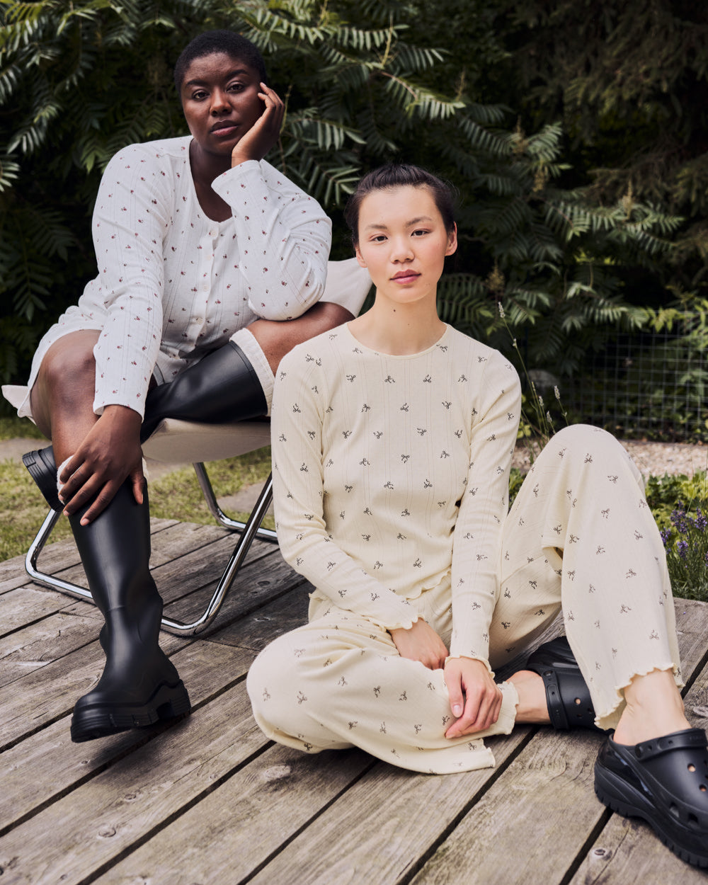 Two women relax outside on a wooden deck with greenery behind them. One lounges cross-legged in jan-n-junes POINTELLE PYJAMA PANTS BUTTERBOW GOTS made from organic cotton, while the other wears a white patterned dress and black boots.