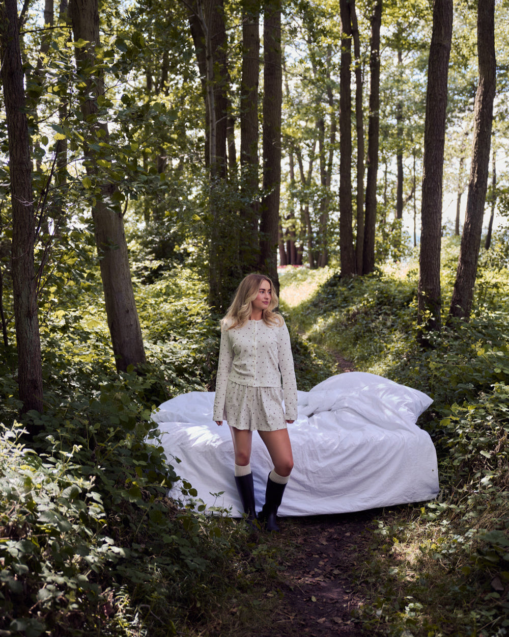 A woman in a light dress and boots stands on a forest path beside a bed covered in white sheets and jan-n-junes POINTELLE PYJAMA SHORTS BLOOMY GOTS, surrounded by tall trees and green foliage in daylight.
