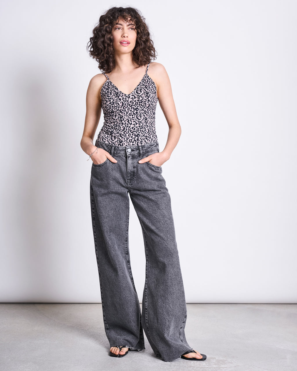 A person with curly hair wears the TOP MATILDA LEO GOTS by jan-n-june— a leo print spaghetti strap top—styled with wide-leg gray organic cotton jeans and black sandals, standing against a plain white background with hands in pockets.