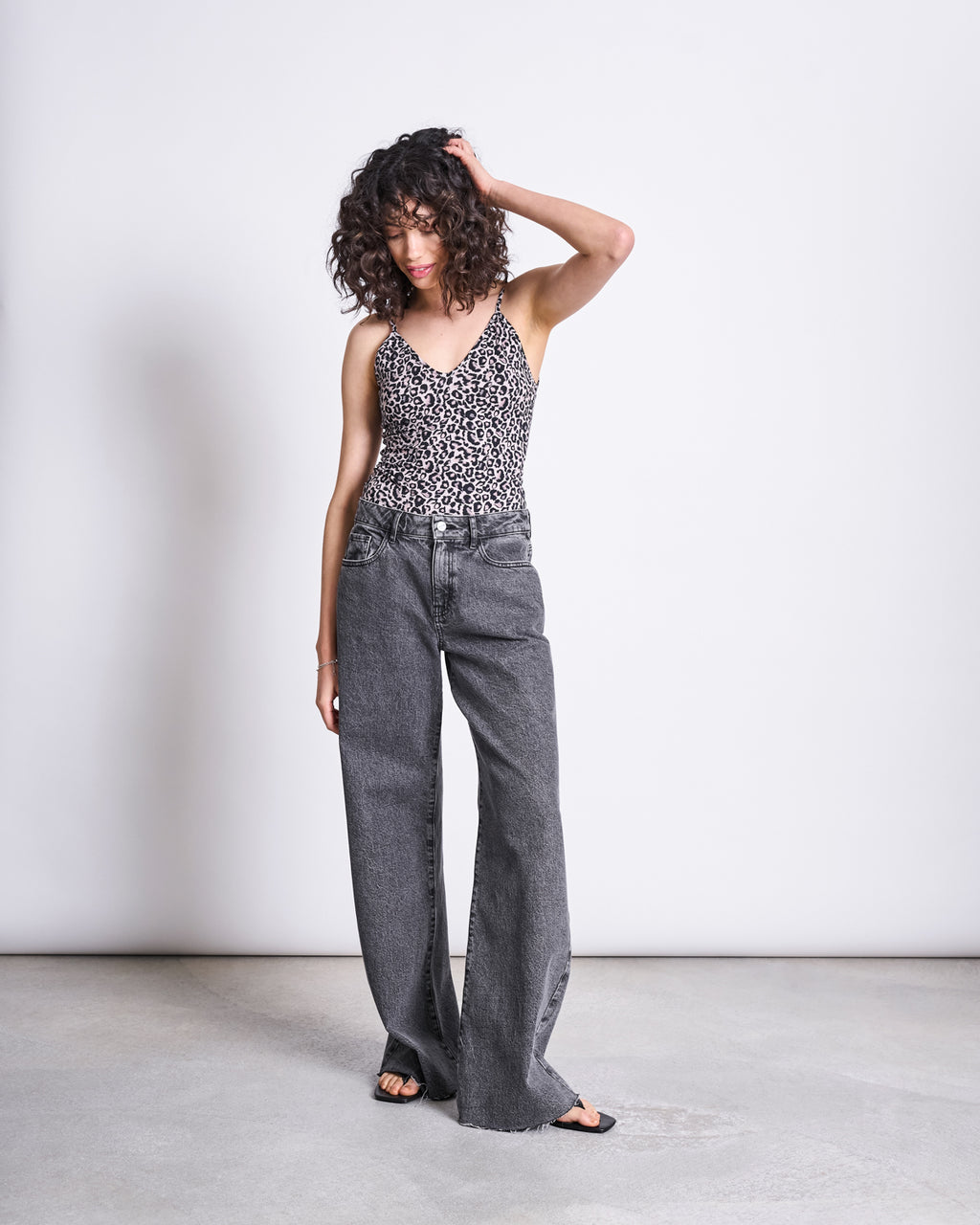 A woman with curly hair wears the jan-n-june TOP MATILDA LEO GOTS with spaghetti straps and loose gray jeans, standing against a plain light background, looking down while touching her head.