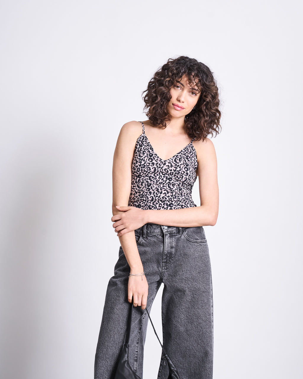 A woman with curly hair wears the jan-n-june TOP MATILDA LEO GOTS, a leo print spaghetti strap top, paired with high-waisted gray jeans. She stands against a plain light background, holding a black bag and crossing her other arm over her body.