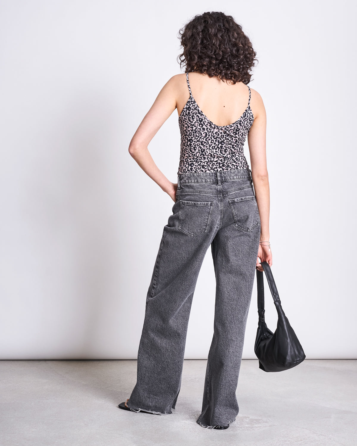 A person with curly hair, wearing the jan-n-june TOP MATILDA LEO GOTS and loose gray jeans, stands facing away from the camera while holding a black handbag in their left hand.