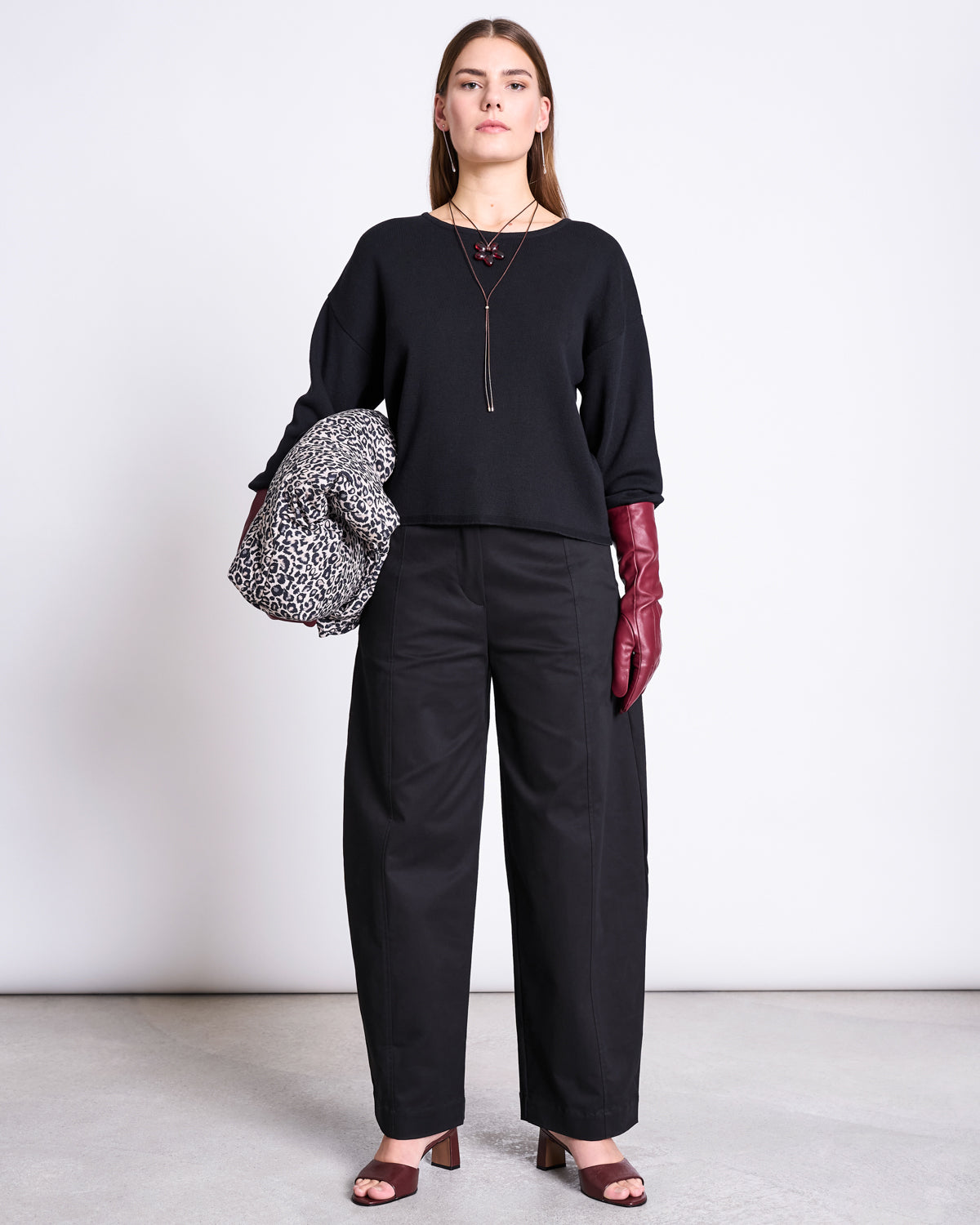 A woman faces forward in a capsule look—black wide-leg pants, jan-n-june’s LIGHT KNIT JUMPER WILLIAMSBURG BLACK GOTS, maroon gloves, brown heeled sandals, long necklace, and holds a leopard print coat against a light backdrop.