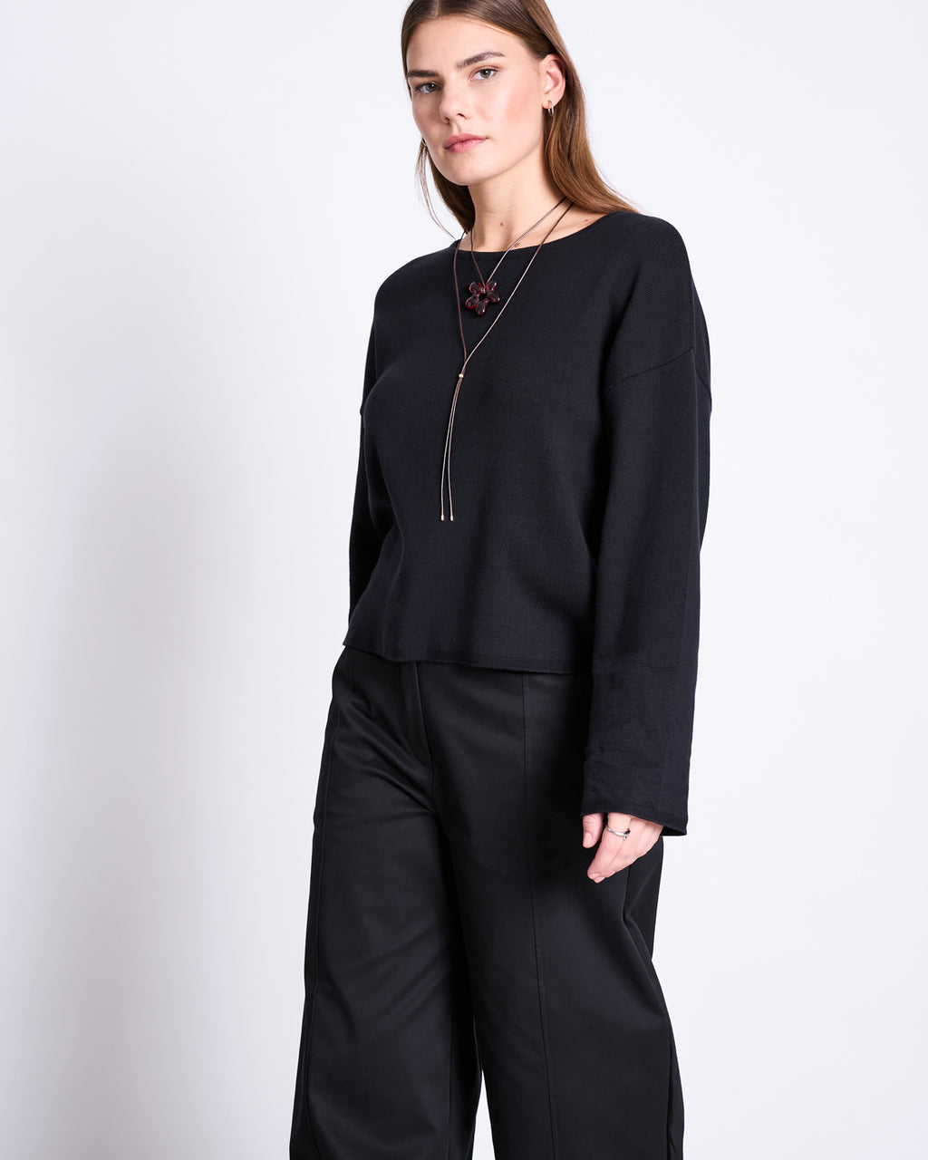 A person with long brown hair wears the jan-n-june LIGHT KNIT JUMPER WILLIAMSBURG BLACK GOTS, paired with wide-leg black pants and a long necklace with a dark pendant, standing against a plain white background.