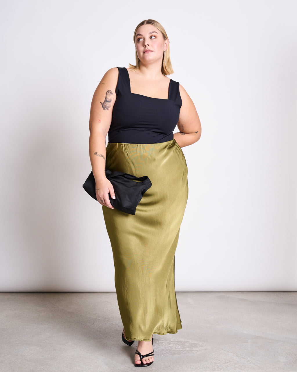 A woman with blonde hair wears the jan-n-june TOP MERIDA BLACK, paired with a shiny olive-green skirt. She holds a black clutch and wears black sandals, standing against a plain light background.