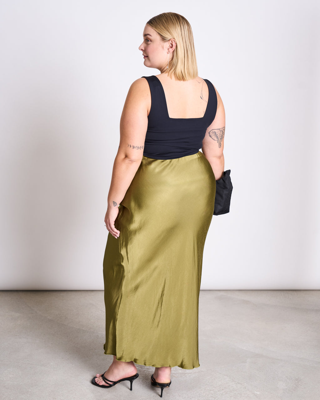A woman with shoulder-length blonde hair and visible tattoos wears the jan-n-june MAXI SKIRT SERIN WOODY OLIVE, a black sleeveless top, black heeled sandals, and holds a small black bag, facing away from the camera.