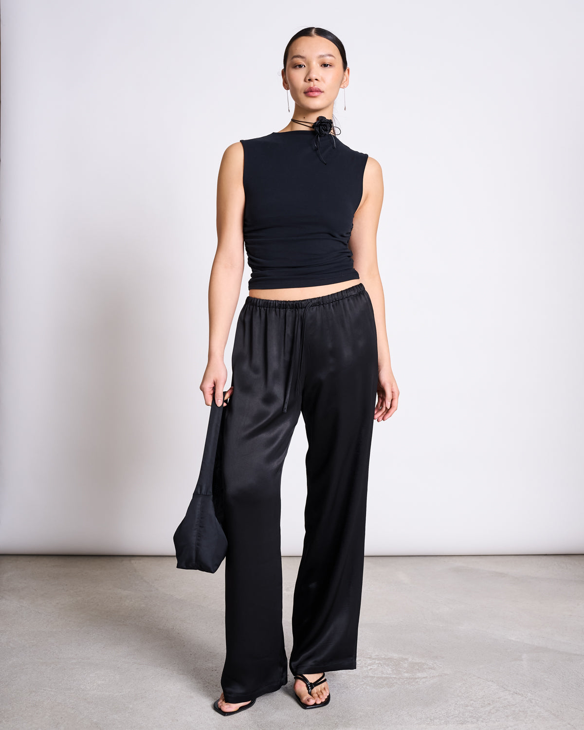 A woman poses indoors against a plain backdrop, wearing jan-n-june’s LOOSE SILKY PANTS FRIS BLACK with a flexible waistband, a sleeveless black top, black sandals, and holding a small black bag as she confidently looks at the camera.