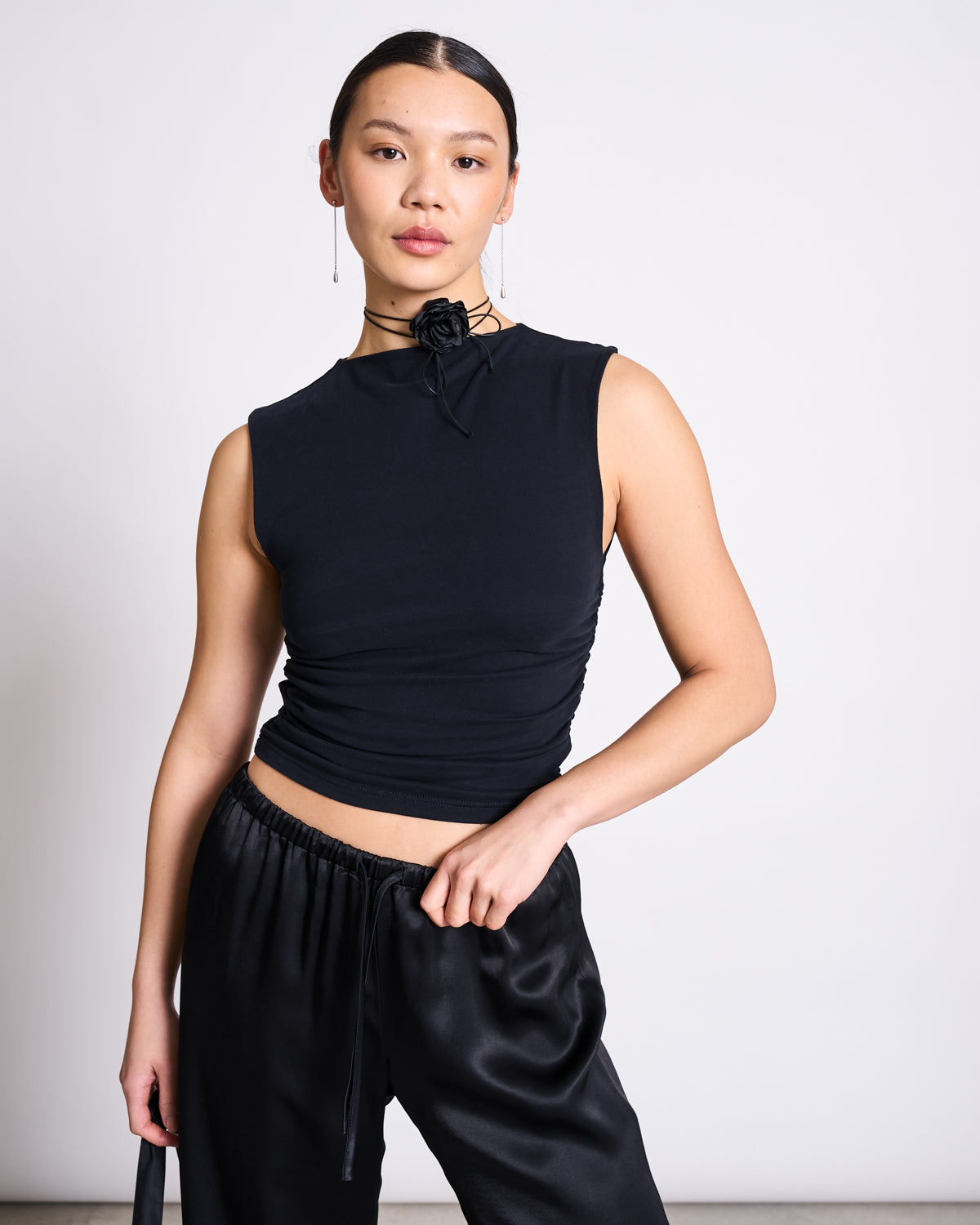 A woman in jan-n-junes LOOSE SILKY PANTS FRIS BLACK pairs the low-waist vegan silk trousers with a sleeveless black top and black rose choker, standing against a white background and gazing confidently at the camera.