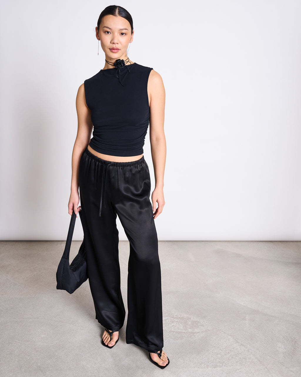 A woman stands against a plain background, wearing the jan-n-june LOOSE SILKY PANTS FRIS BLACK with a sleeveless black top, black flip-flops, and a small black handbag. Her hair is tied back and her expression is neutral.
