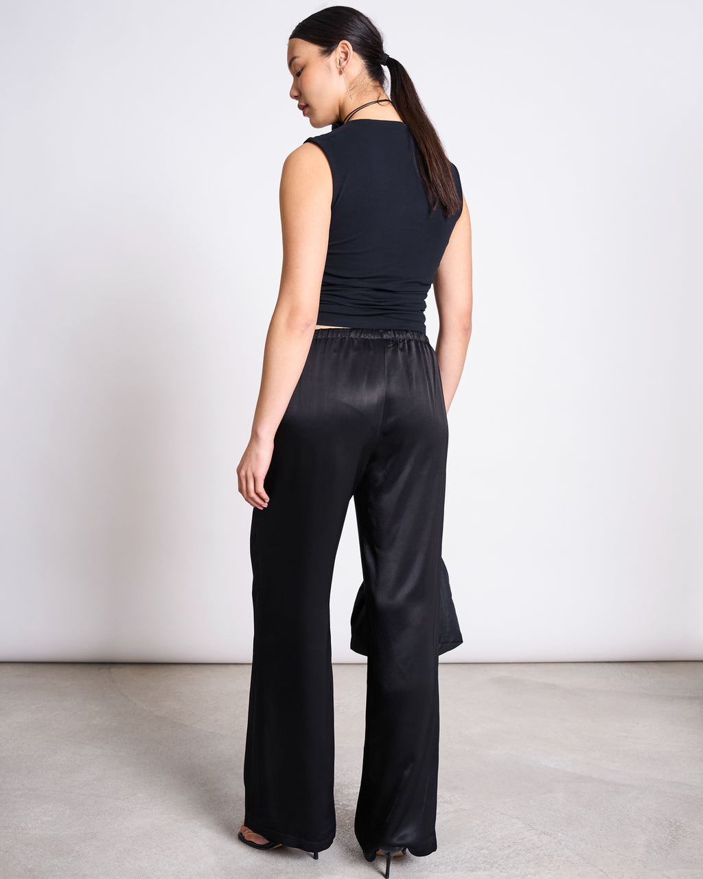 A woman with long dark hair in a low ponytail stands facing away, wearing jan-n-junes LOOSE SILKY PANTS FRIS BLACK styled as wedding pants with a sleeveless black top and heeled shoes, against a minimalist neutral background.