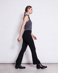 A woman with braided hair walks sideways against a plain white background, wearing a gray sleeveless top and jan-n-june FLARED PANTS MIOSA BLACK GOTS with an elastic waistband, paired with black boots.