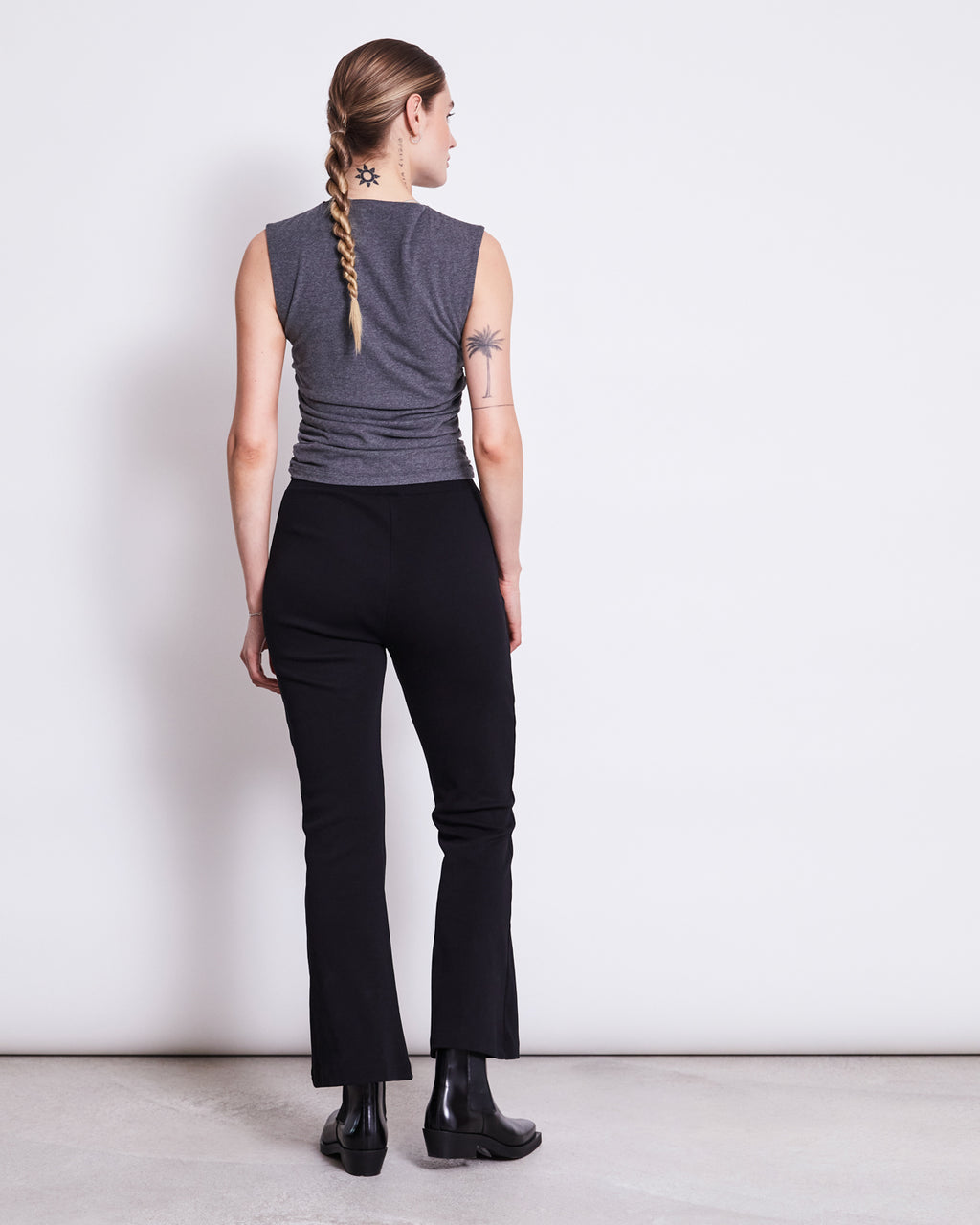 A person with a long braid and tattoos wears a sleeveless gray top, jan-n-junes FLARED PANTS MIOSA BLACK GOTS with an elasticated waistband, and black boots, standing against a plain white wall.