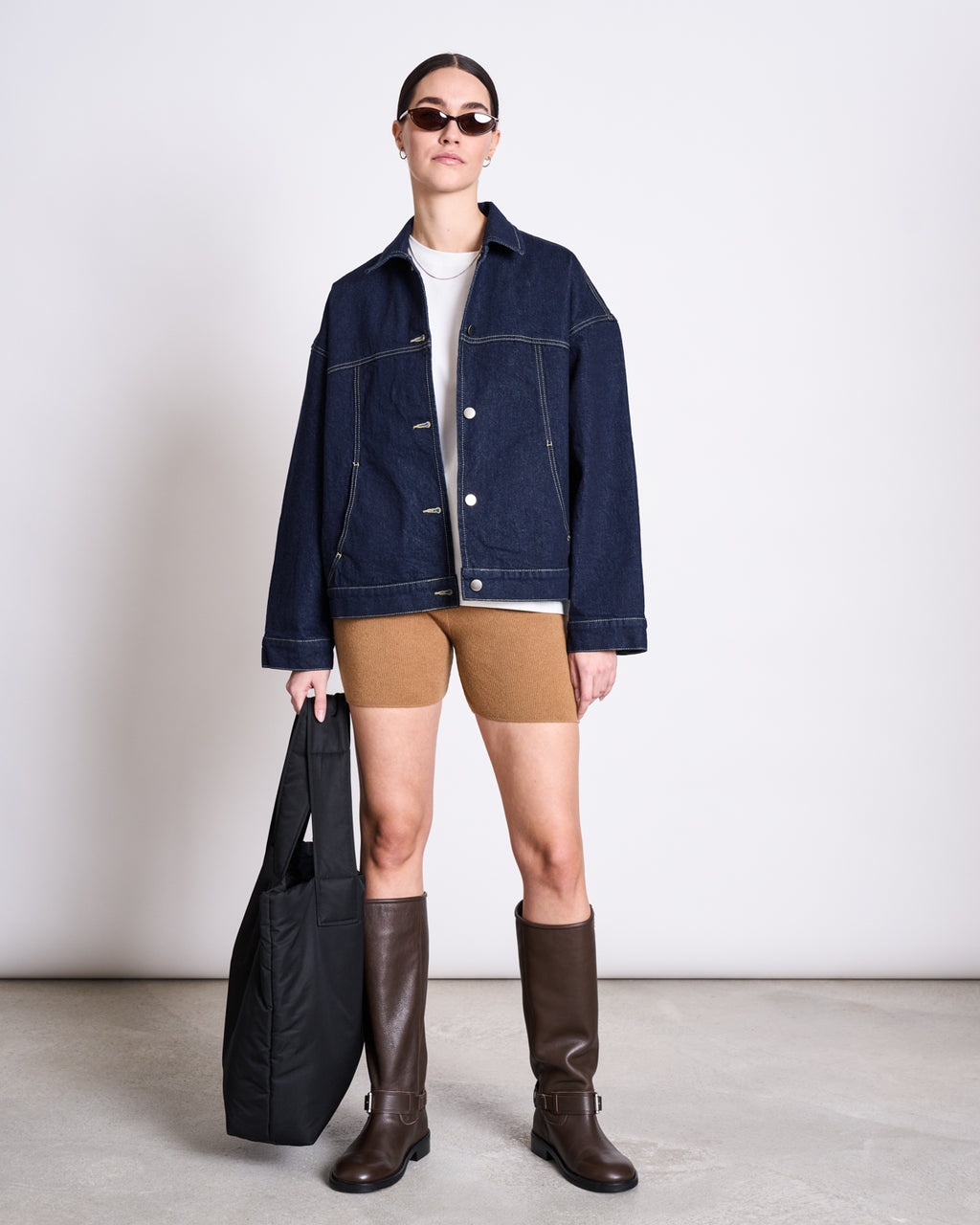 A woman stands against a plain background wearing dark sunglasses, a dark denim jacket from the HANDLE WITH CARE collection, a white T-shirt, jan-n-junes BIKER SHORTS ALVA WOOD, knee-high brown boots, and holding a large black tote bag.
