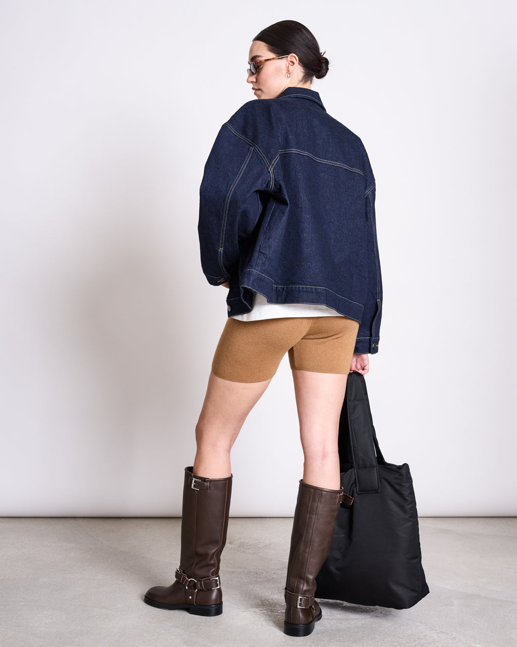A woman faces away, wearing BIKER SHORTS ALVA WOOD by jan-n-june, an oversized denim jacket, tall brown boots, sunglasses, and holding a large black tote against a plain light background.