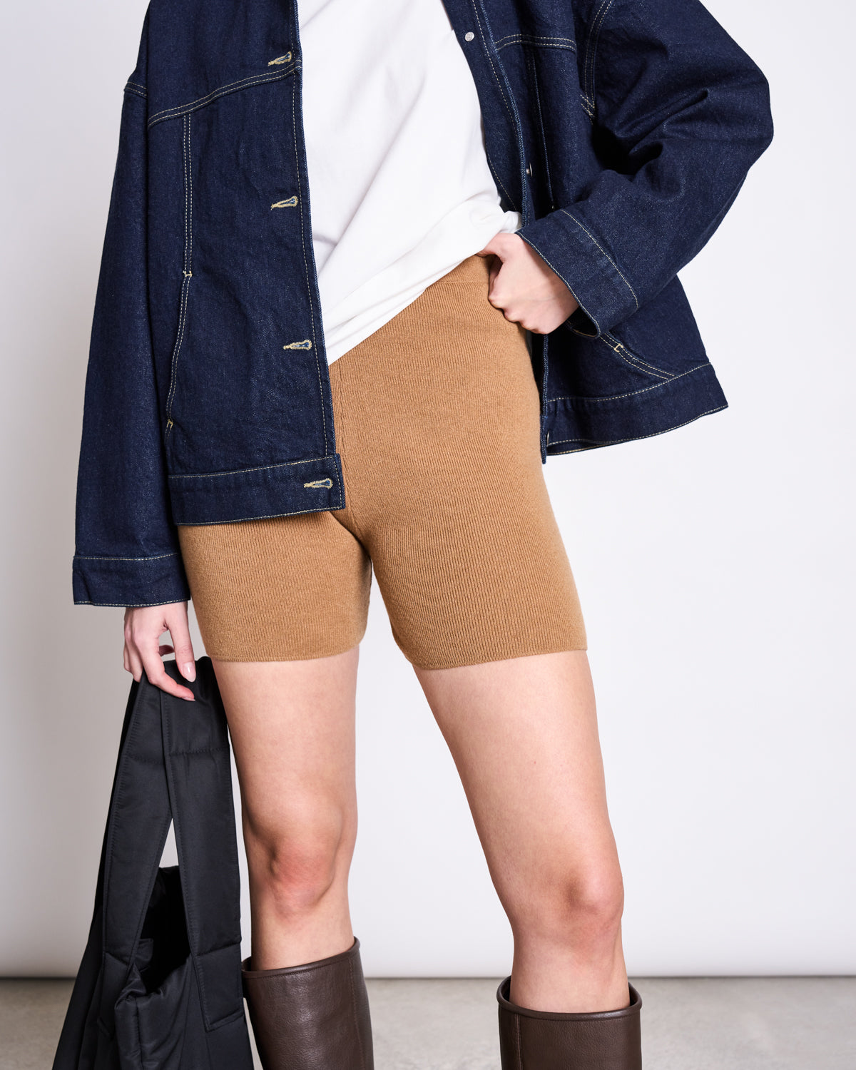 Person wears a dark denim jacket, white shirt, BIKER SHORTS ALVA WOOD by jan-n-june, and brown knee-high boots from the HANDLE WITH CARE collection by ALVA, holding a black bag against a plain light background.