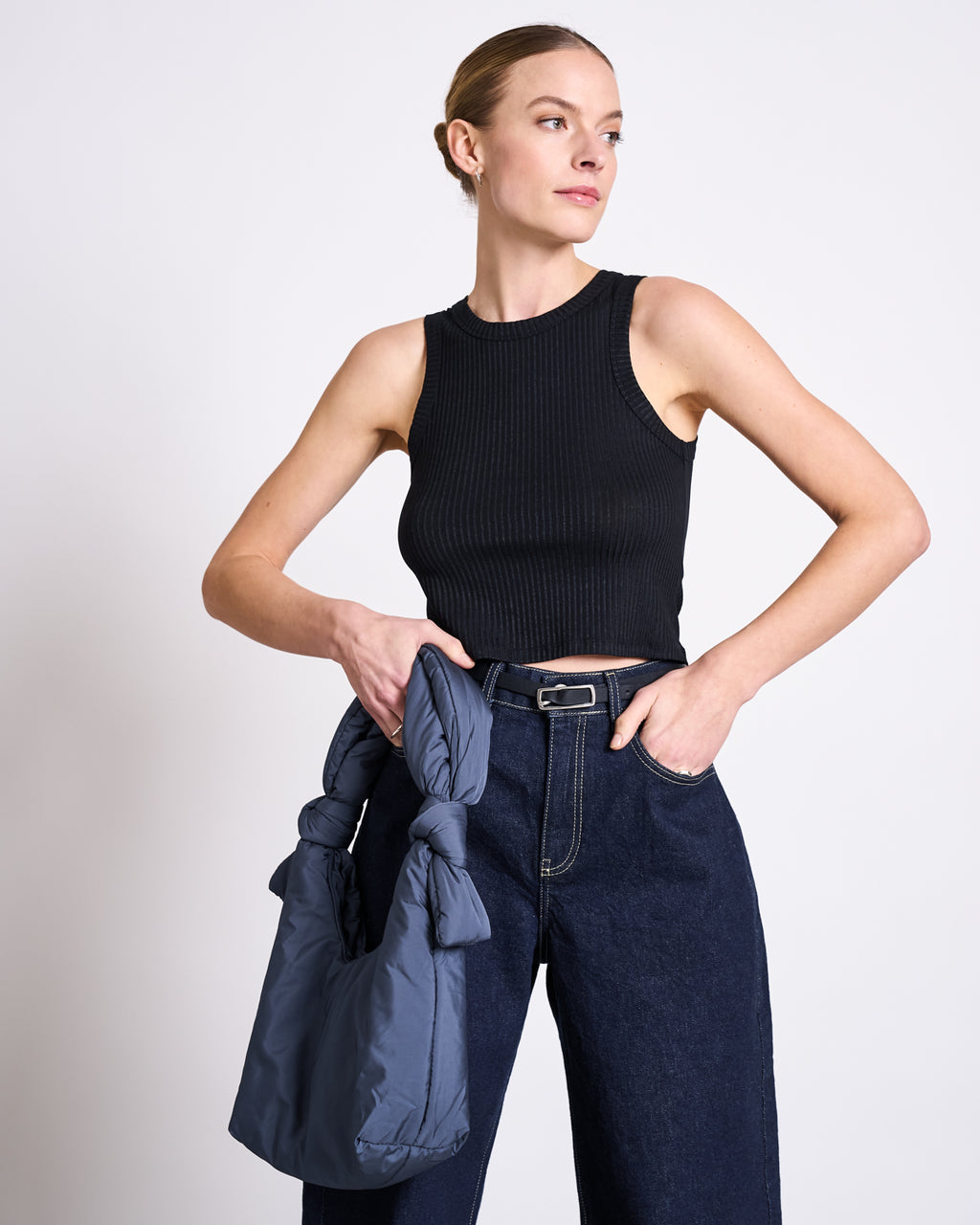A woman in the jan-n-june LYOCELL RIB TANK TOP PORTO BLACK and high-waisted dark jeans holds a gray puffy handbag, her hair pulled back, looking off to the side against a plain background.