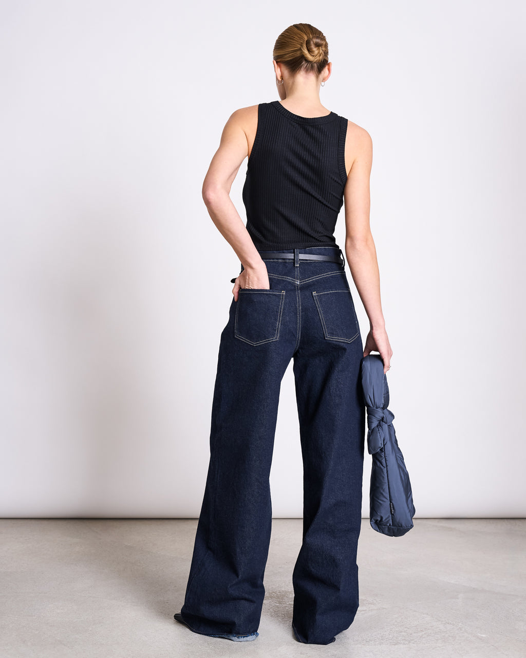 A person with blonde hair in a bun wears the jan-n-june LYOCELL RIB TANK TOP PORTO BLACK and wide-leg dark blue jeans, standing with their back to the camera and holding a rolled-up navy jacket in their left hand.