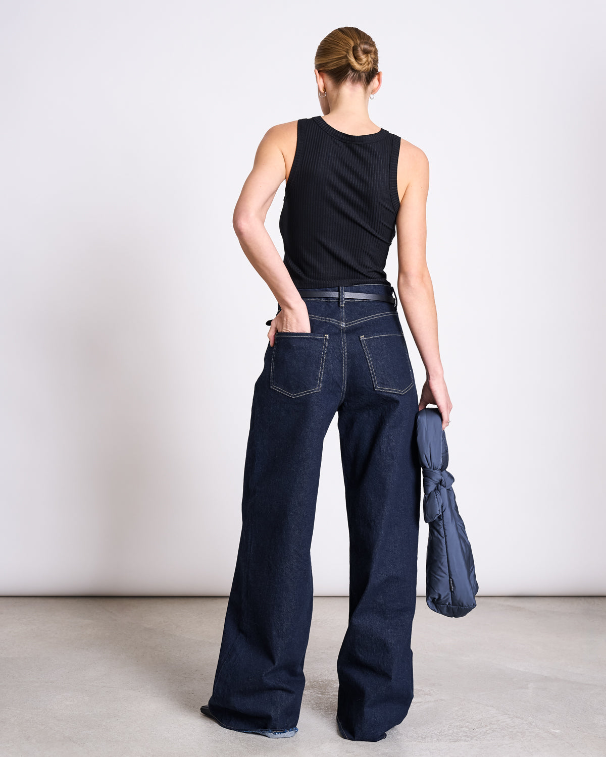 A person with blonde hair in a bun wears the jan-n-june LYOCELL RIB TANK TOP PORTO BLACK and wide-leg dark blue jeans, standing with their back to the camera and holding a rolled-up navy jacket in their left hand.