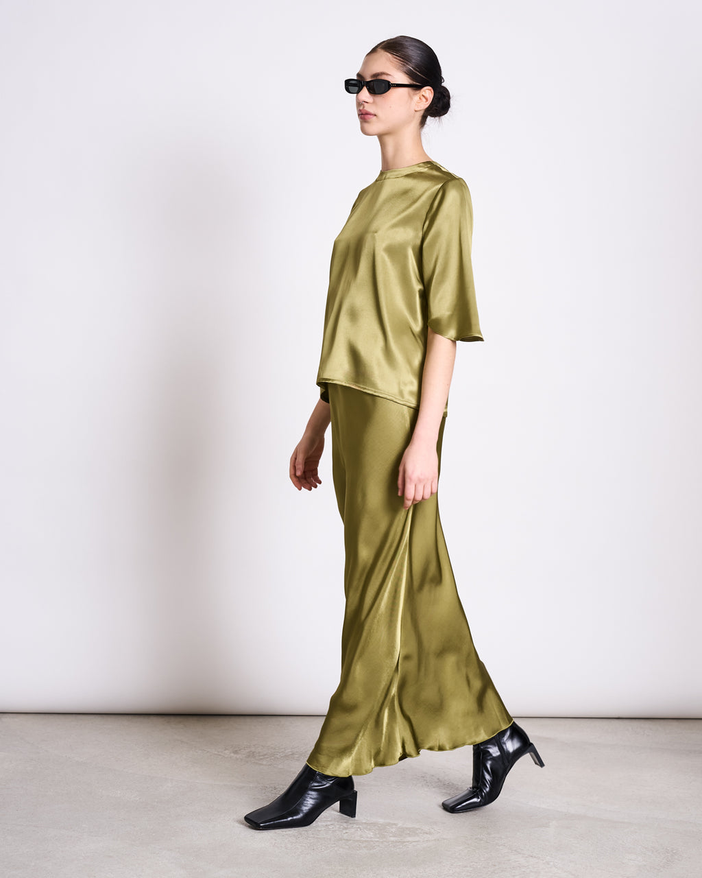A woman in sunglasses stands in profile against a light background, wearing jan-n-junes SILKY SHIRT NAMORA WOODY OLIVE and matching long ENKA® Viscose skirt, paired with black heeled boots.