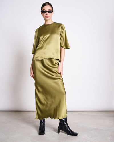 A woman in a loose-fitting jan-n-june SILKY SHIRT NAMORA WOODY OLIVE with a matching long skirt, black ankle boots, and dark sunglasses stands against a plain white background.